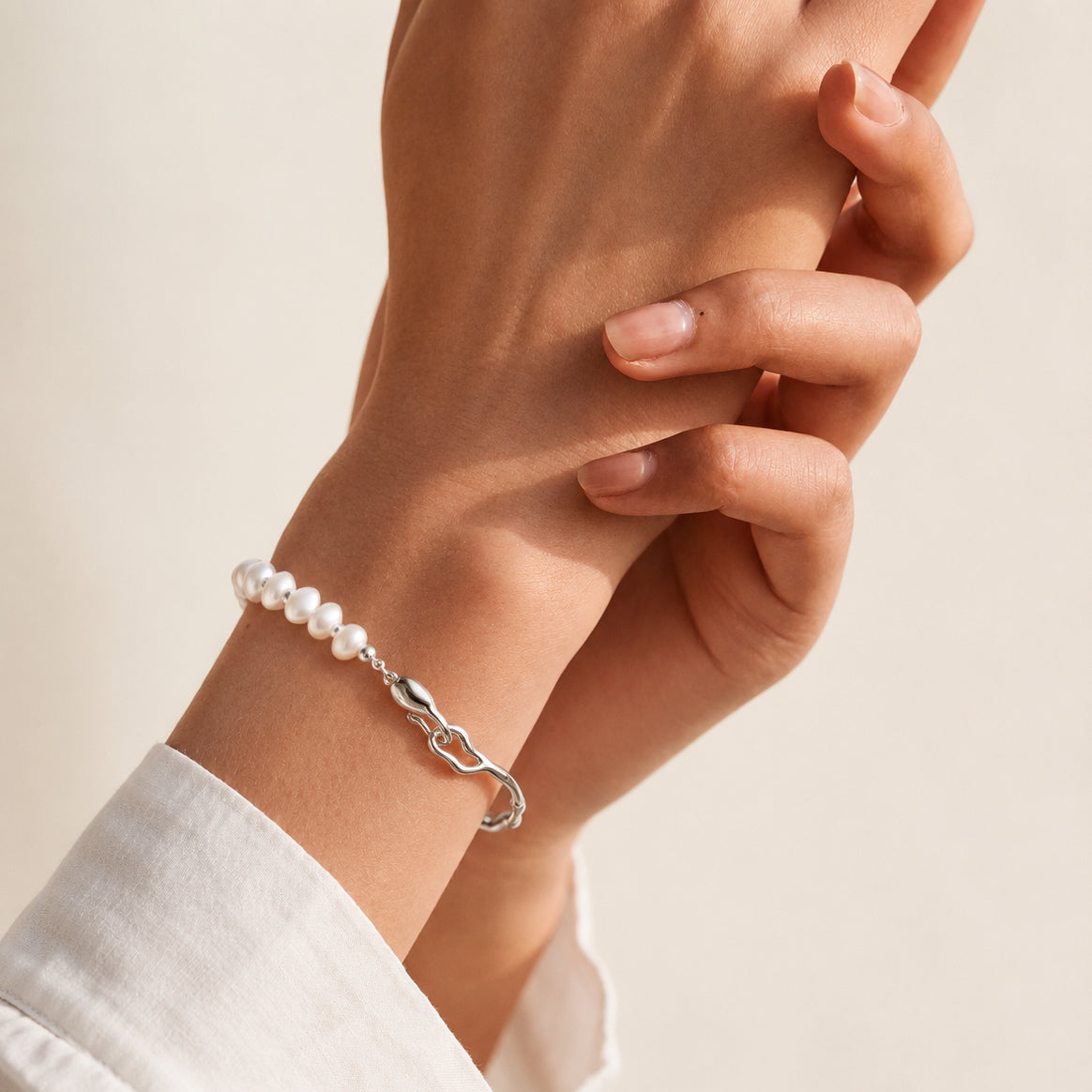 A close-up of a person's wrist wearing a delicate bracelet made of white pearls and a polished silver clasp, paired with a white collared shirt against a soft beige background.