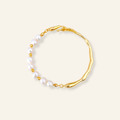 A gold-toned bracelet featuring a wavy, organic-shaped band and a row of white freshwater pearls separated by small gold beads, set against a solid cream background.
