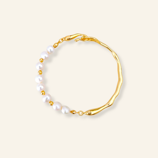 A gold-toned bracelet featuring a wavy, organic-shaped band and a row of white freshwater pearls separated by small gold beads, set against a solid cream background.