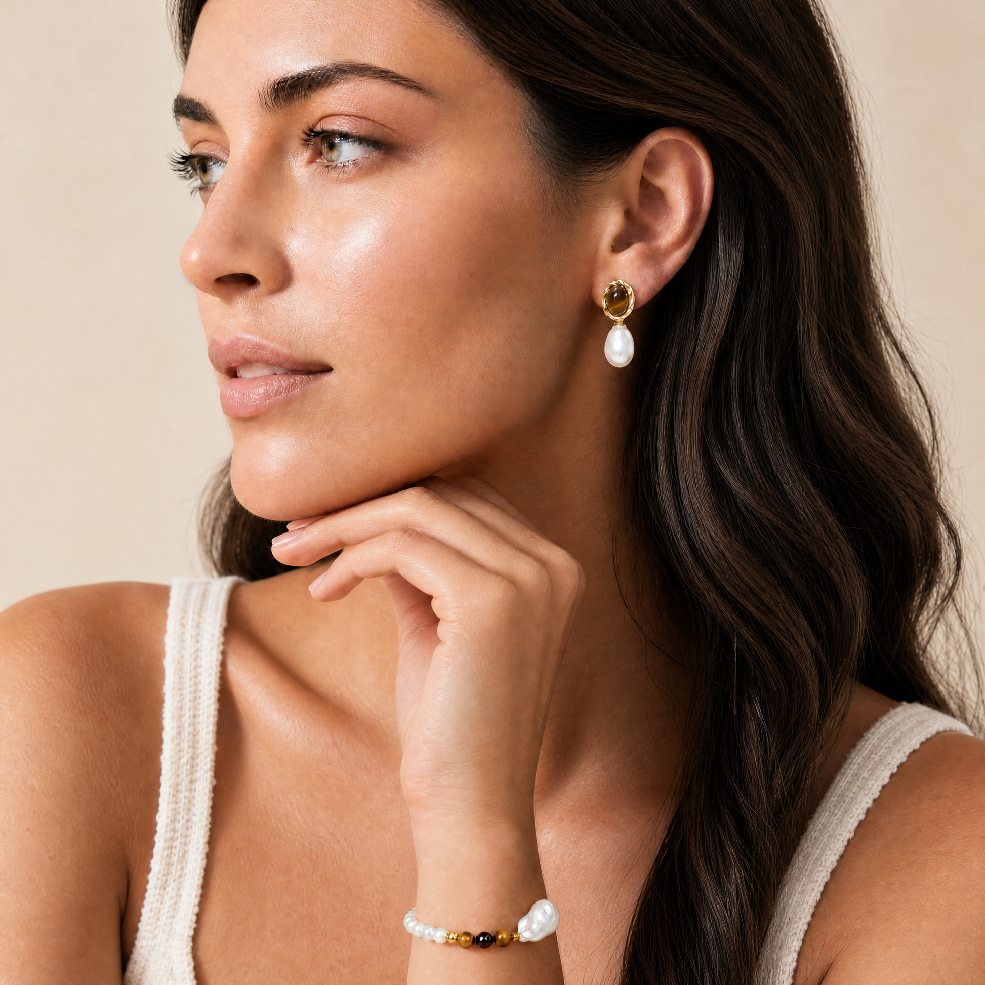 A close-up lifestyle shot of a woman wearing a pearl earring and a pearl bracelet, set against a warm beige background.