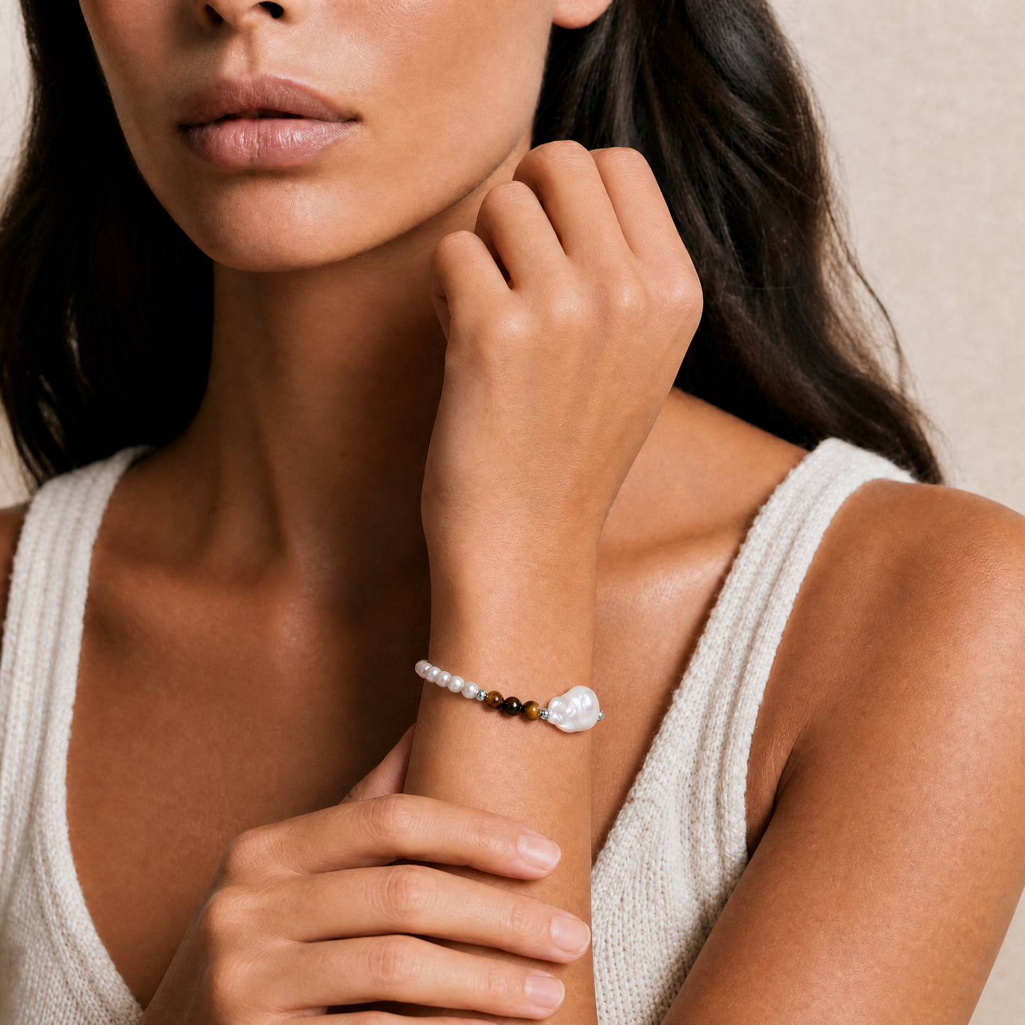 A close-up lifestyle shot of a model wearing a delicate beaded bracelet on her wrist. The bracelet features small white pearls, a dark brown tiger eye stone