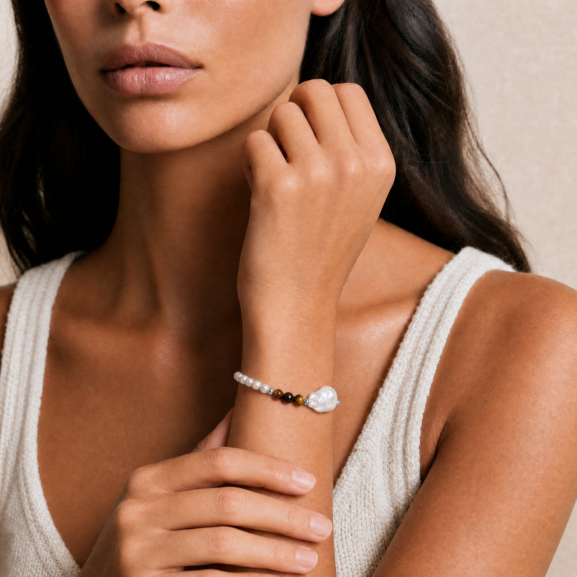 A close-up lifestyle shot of a model wearing a delicate beaded bracelet on her wrist. The bracelet features small white pearls, a dark brown tiger eye stone