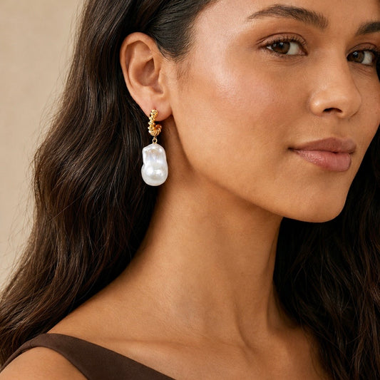 A close-up of a woman wearing a gold huggie hoop earring with a large white baroque pearl drop.