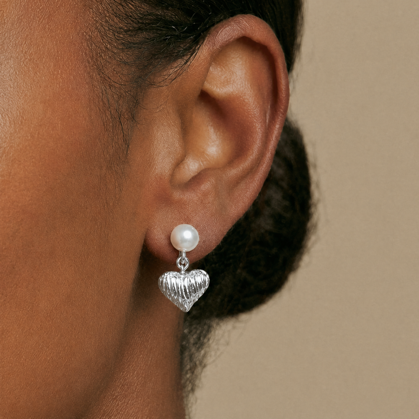 Close-up of an ear wearing a silver heart-shaped earring with a pearl detail on a beige background